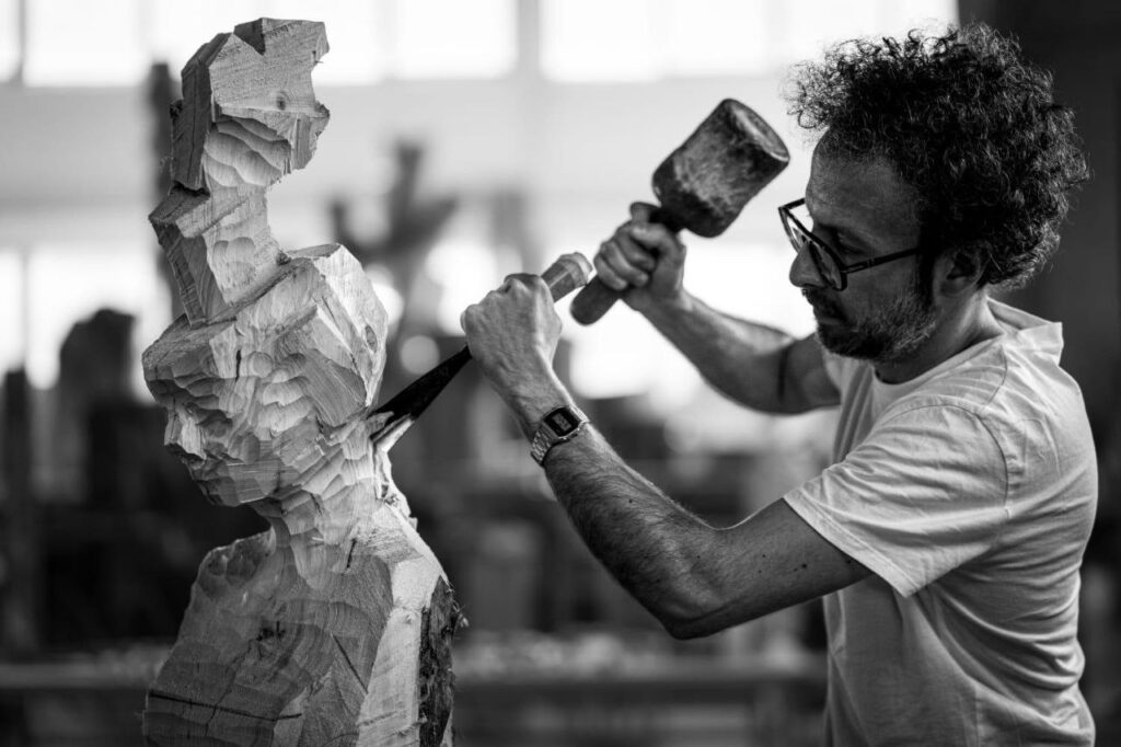 The image depicts the sculptor Arcangelo Ambrosi intent on carving a wooden sculpture on a solid workbench; captured in the moment when the mallet strikes the gouge and begins to carve into the wood.
